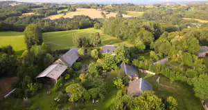 Luftaufnahme eines Campinghofs mit mehreren Gebäuden, Gartenwegen und Bäumen inmitten grüner Felder und Landschaft.