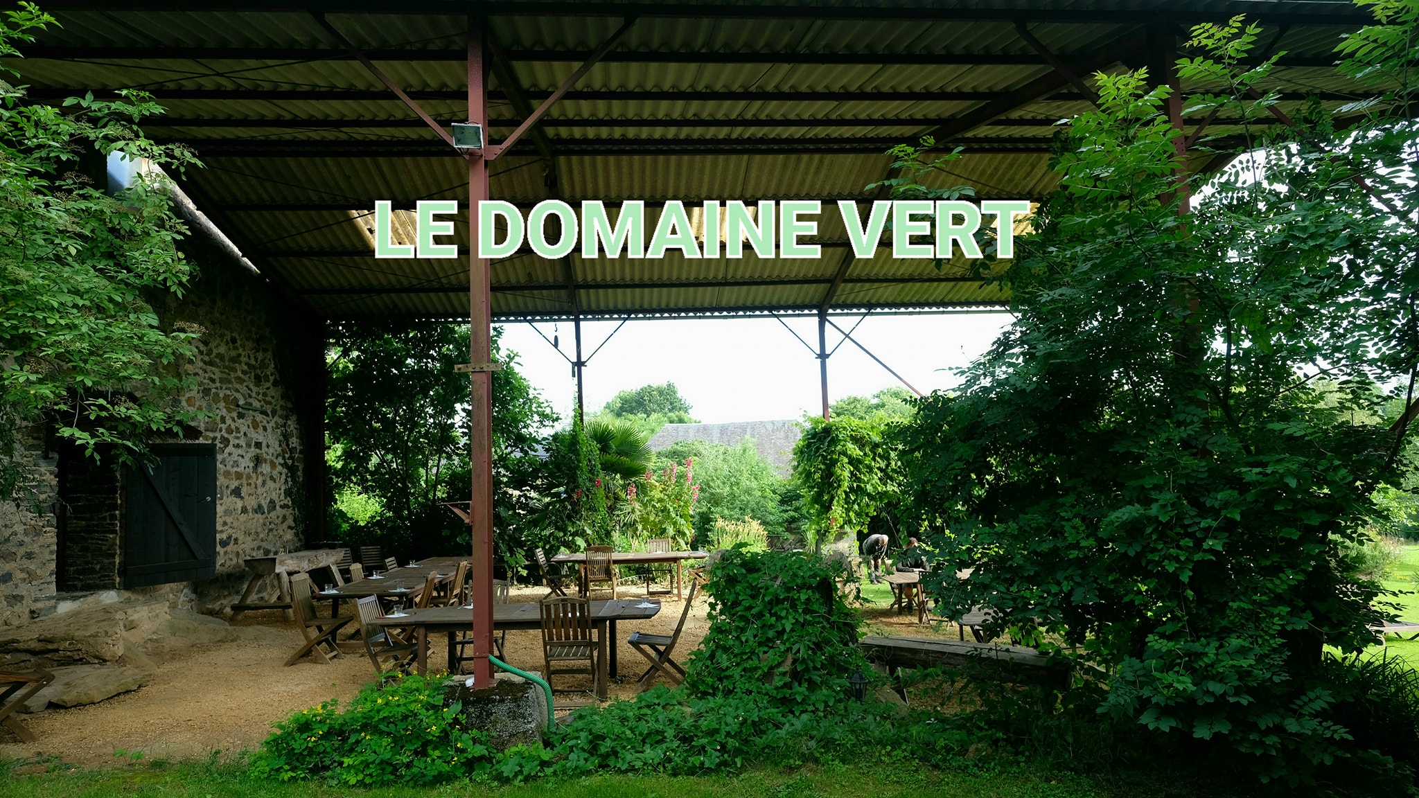 Terrasse am Campingplatz mit Holzdach, Sitzgruppe und Blick ins Grüne, Schild „LE DOMAINE VERT“ im Vordergrund.
