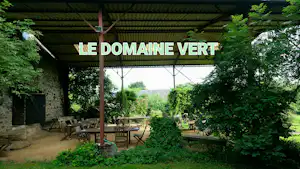 Terrasse am Campingplatz mit Holzdach, Sitzgruppe und Blick ins Grüne, Schild „LE DOMAINE VERT“ im Vordergrund.