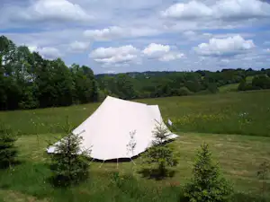 Weißes Campingzelt auf einer grünen Wiese, umgeben von Büschen und Bäumen, im Hintergrund ein bewaldetes Tal.