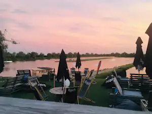 Campingstühle und Sonnenschirme auf einer Terrasse am Flussufer, im Hintergrund ruhiges Wasser und ein grüner Uferstreifen.