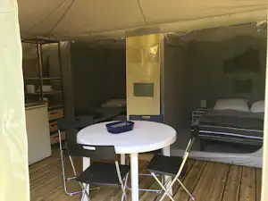 Innenansicht eines Campingzelts mit rundem Tisch und Stühlen, daneben Betten und Holzfußboden.