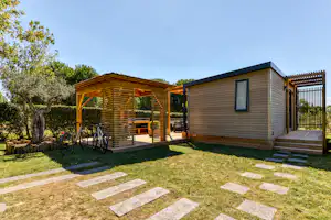 Holz-Ferienhaus mit Terrasse, Pergola und Fahrrädern am Campingplatz, umgeben von Hecken und Rasen.
