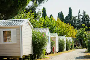 Reihe von Mobilheimen mit weißen Fassaden und Schiebeläden, umgeben von Büschen, Bäumen und einem Kiesweg im Campingbereich.