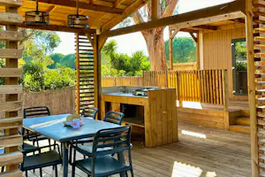 Holzterrasse mit Esstisch und Stühlen, Grill an der Küchenzeile, Holzgeländer und Blick in den Garten mit Bäumen.