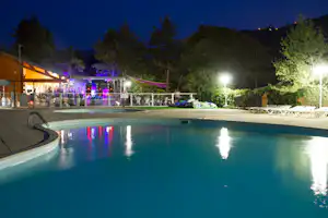 Beleuchteter Campingpool am Abend mit Liegen am Rand, Zaun und Bäumen im Hintergrund.