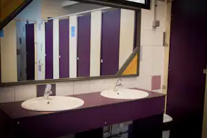 Waschbecken im Sanitärraum mit zwei Spülbecken, Armaturen und Spiegel, dahinter mehrere Kabinen mit lila Türen.