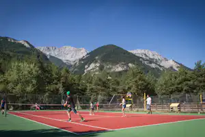 Tennisplatz mit Spielern, rotem Boden und Netz, im Hintergrund Wald und schroffe Berge unter blauem Himmel.