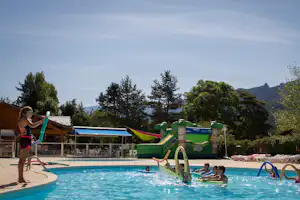 Kinder am Pool mit Wasserrutsche und Hüpfburg im Campingbereich, im Hintergrund Bäume und Berge.