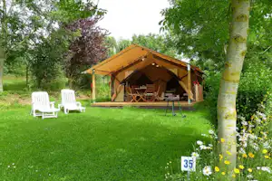 Glamping-Zelt mit Holzpodest und Sitzbereich, zwei weiße Liegestühle auf dem Rasen, umgeben von Bäumen und Blumen.