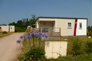 Mobilheim mit Holzterrasse, Stühlen und Blumenbeeten am Weg, daneben ein weiteres Mobilheim im Campinggelände.