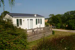Ferienhaus mit Terrasse, Holzgeländer und grünen Türen, umgeben von Büschen und Bäumen auf dem Campingplatz.