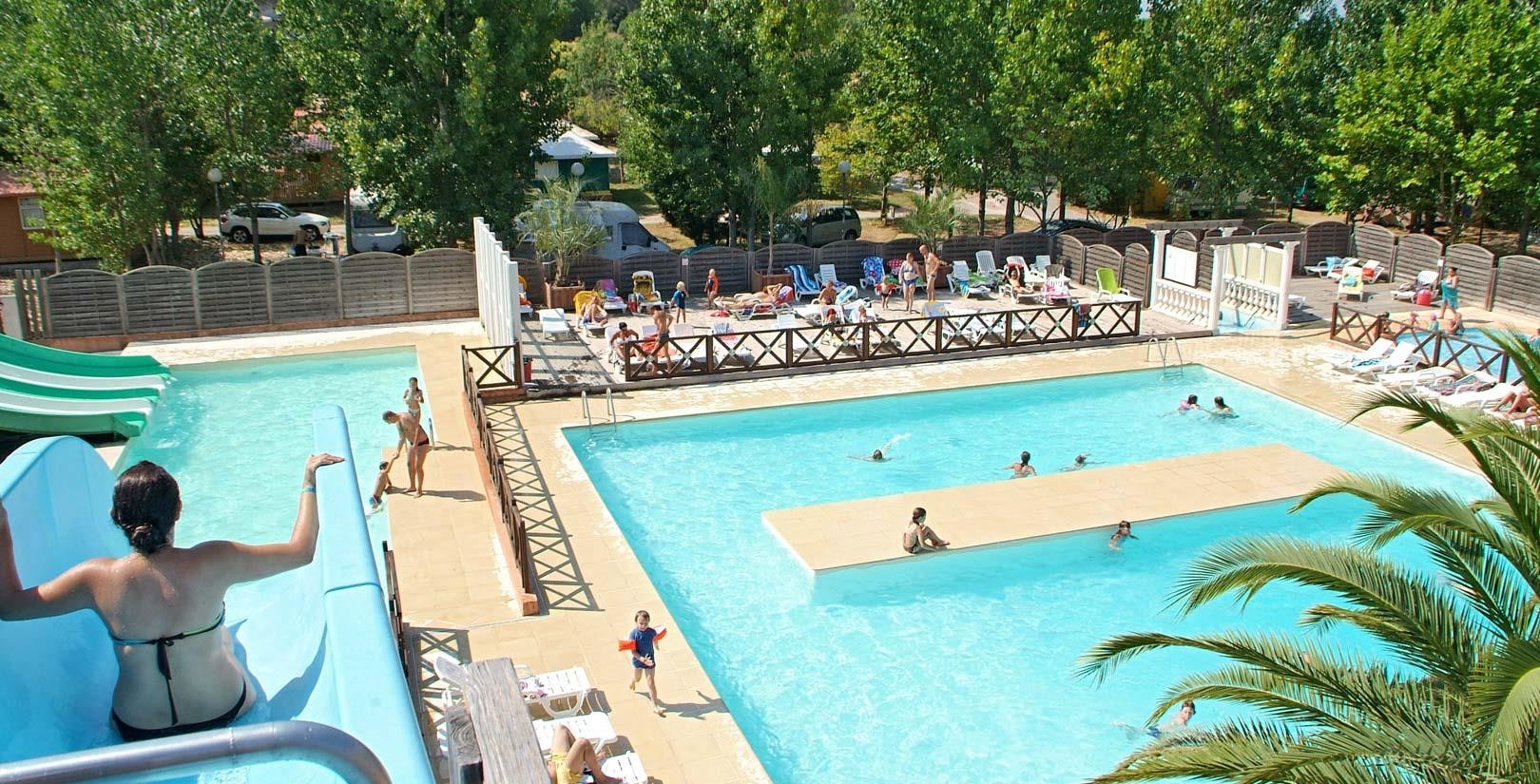 Großes Camping-Schwimmbecken mit Liegen, Palmen und Zaun, daneben ein weiteres Becken und Wege im Hintergrund.