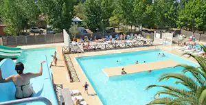 Großes Camping-Schwimmbecken mit Liegen, Palmen und Zaun, daneben ein weiteres Becken und Wege im Hintergrund.