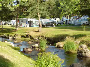 Campingplatz am Bach mit Wohnwagen und Zelten, grüner Wiese und Bäumen am Ufer.
