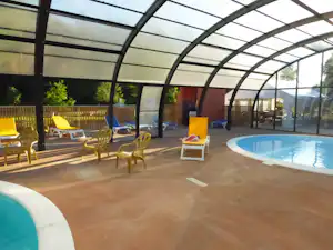 Innenbereich mit beheiztem Pool, Liegestühlen und Glasdach, daneben gelbe Sonnenliegen und Sicht auf den Außenbereich.