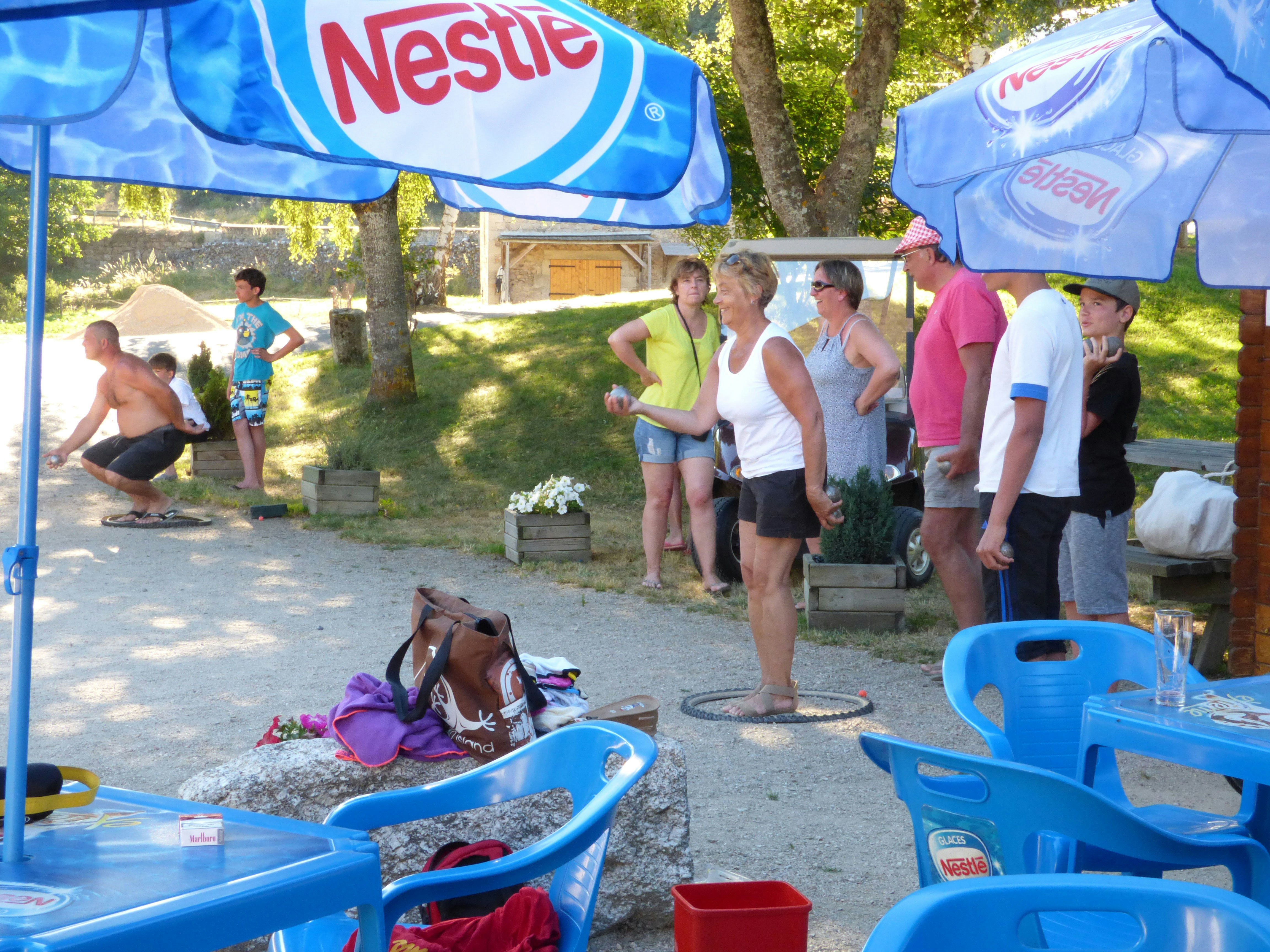 Campingplatz mit Kindern und Erwachsenen am Weg, blaue Planschbecken unter Sonnenschirmen, Bäume und Holzhäuschen im Hintergrund.