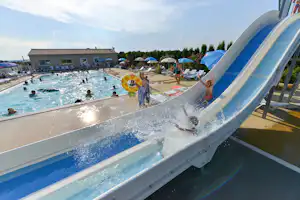 Wasserrutsche am Campingpool, daneben Liegen und ein Becken mit mehreren Schwimmern vor dem Gebäude.