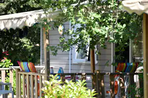 Holzterrasse mit bunten Gartenstühlen und Sonnenschirm vor einem Wohnwagen, umgeben von grünen Bäumen und Pflanzen.