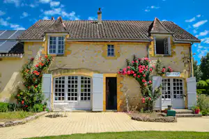 Rezeption eines Campinghauses mit Steinfassade, weißen Fenstern und Türen, Blumenbeeten und gepflastertem Eingang unter blauem Himmel.