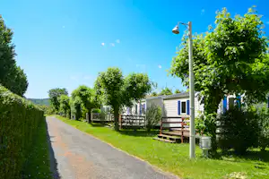Wohnwagen am Wegesrand, Holzterrasse und Bank, umgeben von Bäumen und grünem Rasen unter blauem Himmel.