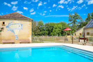 Pool am Campingplatz mit Terrasse, Holzgeländer und Gartenbäumen vor einem hellen Himmel.