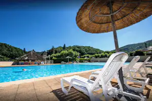 Weiße Liegen am Pool mit Sonnenschirm, im Hintergrund Bungalows und Hecken auf dem Campingplatz.