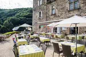 Terrasse mit vielen gedeckten Tischen und Sonnenschirmen vor einem Steingebäude, im Hintergrund Waldhügel.