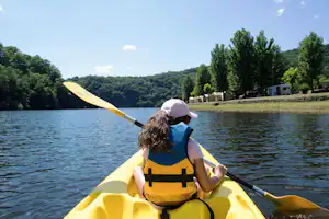 Person im gelben Kajak mit Schwimmweste paddelt auf dem See, im Hintergrund Ufer mit Bäumen und Wohnwagen.