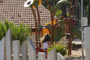 Buntes Klettergerüst auf dem Campingplatz, mit Rutsche, Holzpfosten und Blick auf den gepflasterten Weg und Bäume.