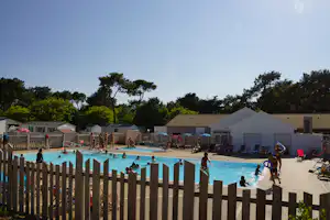 Schwimmbad auf dem Campingplatz mit Liegestühlen, Wohnwagen und Zelten hinter einem Holzzaun.