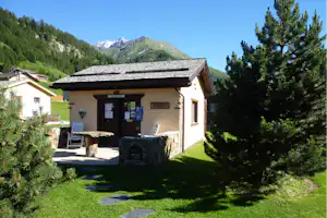Rezeption eines Campingplatzes in einer kleinen Hütte mit Sitzbank, Infotafeln und Blick auf grüne Berge und Fichten.