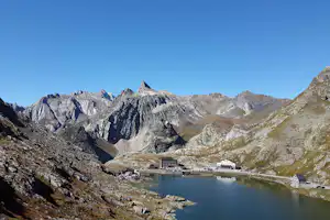 Bergsee im Tal mit Blick auf schroffe Alpenfelsen und mehrere Gebäude am Ufer.