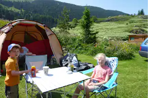 Campingzelt auf einer Wiese am Berg, daneben Tisch und Stühle, im Hintergrund Bäume und ein Holzhaus.