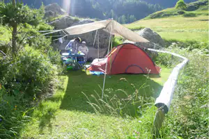 Campingzelt mit Vorzelt und Tisch auf einer Wiese, im Hintergrund Felsen und bewaldete Berge.