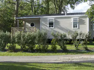 Graues Mobilheim mit Holzveranda, Fenster und Eingangstür, umgeben von Büschen und Bäumen im Grünen.