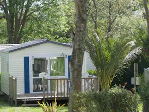 Mobilheim mit Veranda und weißen Geländern, umgeben von Bäumen und Sträuchern im Campingbereich.