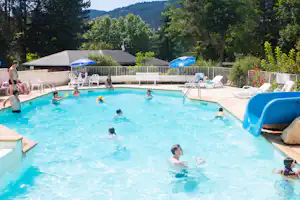 Campingpool mit blauem Wasser, Rutsche, Liegestühlen und Sonnenschirmen; Gäste planschen im Becken vor dem Gebäude im Grünen.