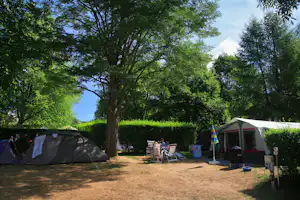 Zelte auf einem Campingplatz unter großen Bäumen, mit Gartenstühlen und Tisch auf dem Stellplatz.