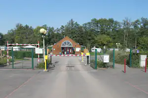 Eingang eines Campingplatzes mit Schranke, Holzgebäude mit rundem Fenster und Zaun, im Hintergrund Bäume.