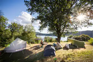 Campingzelt und weitere Zelte auf einem Stellplatz am See, mit Bäumen, Rasen und Blick auf das Wasser.