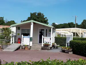 Rezeption am Campingplatz-Gebäude mit Eingang, Infotafeln und Sitzbank vor dem Gebäude, umgeben von Hecken und Bäumen.