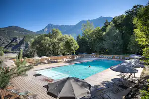 Schwimmbecken auf einer Holzterrasse mit Liegen und Sonnenschirmen, umgeben von Bäumen und Bergen im Hintergrund.