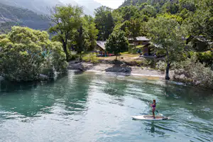 Stand-up-Paddle auf dem Fluss vor einem Campingplatz mit Stellplätzen, Bäumen und kleinen Gebäuden im Tal.