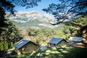 Campingzelte mit Markisen auf einem Stellplatz im Wald, im Hintergrund hohe Alpenberge und grünes Tal.