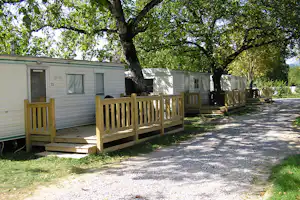 Wohnwagen mit Holzterrasse und Eingangstür, umgeben von Bäumen, entlang eines Kieswegs auf dem Campingplatz.