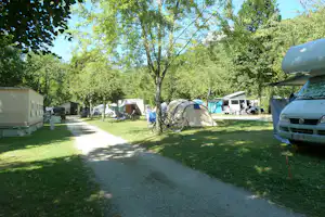 Campingplatz mit Zelt, Fahrrädern und Autos am Weg, umgeben von Rasen und hohen Bäumen.