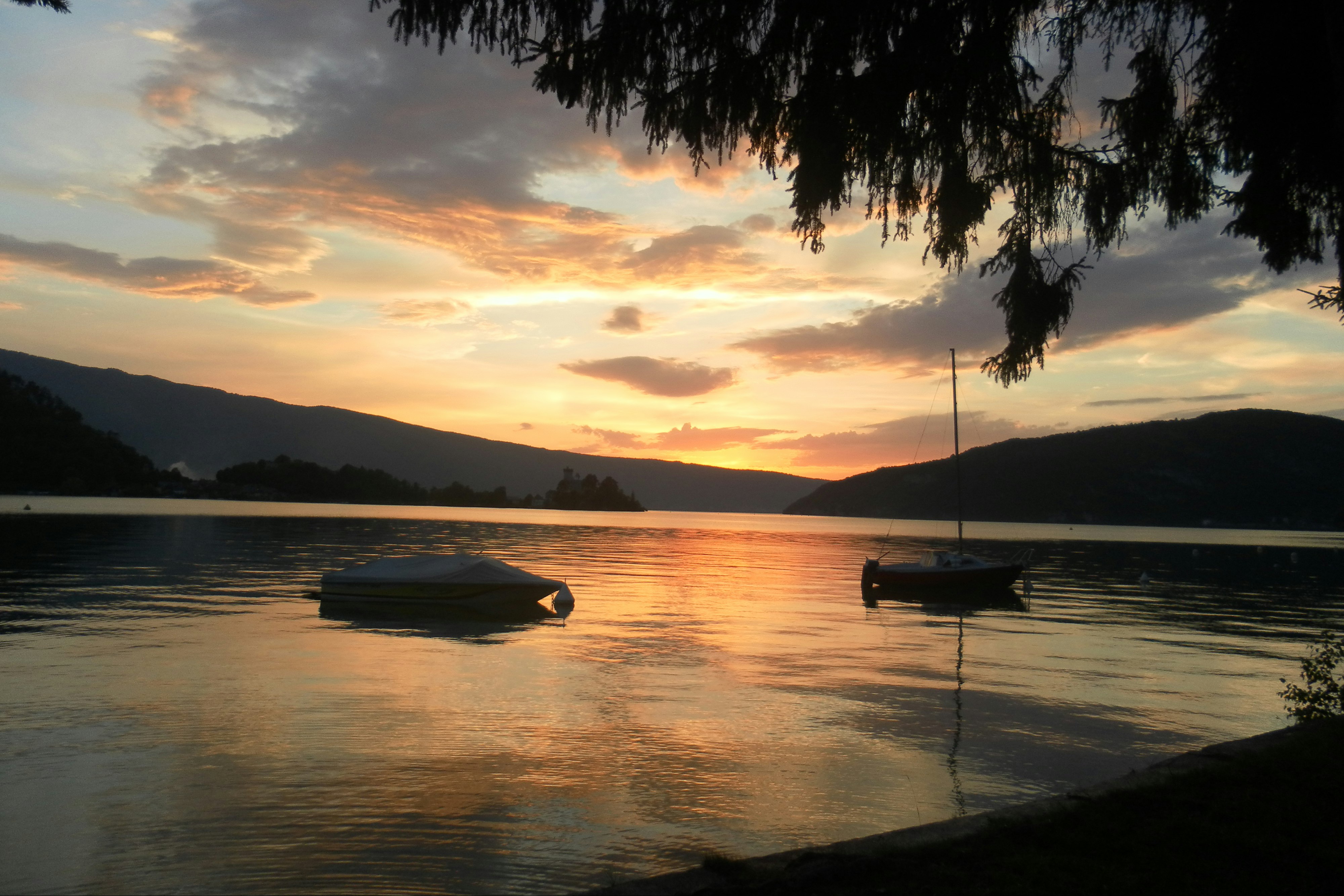 Ruderboot und kleines Boot am Seeufer bei Sonnenuntergang, im Vordergrund Wasser und Sandstrand, im Hintergrund Hügel.