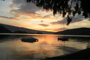 Ruderboot und kleines Boot am Seeufer bei Sonnenuntergang, im Vordergrund Wasser und Sandstrand, im Hintergrund Hügel.