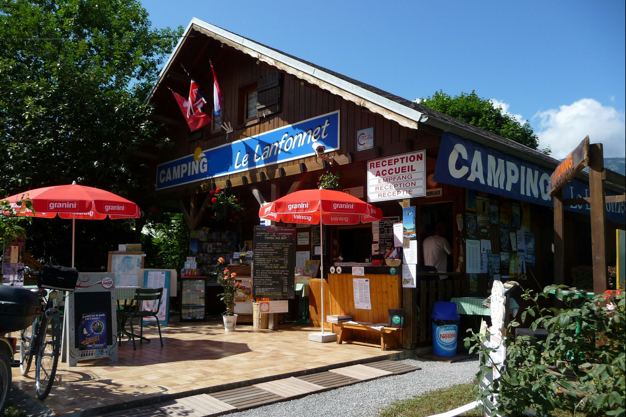 Camping-Rezeption in einem Holzgebäude mit Schildern, roten Marktschirmen und Fahrrad vor dem Eingangsbereich.